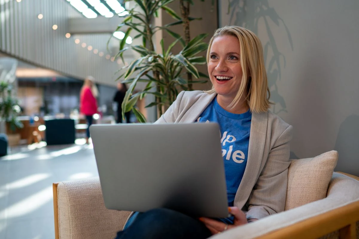 A smiling person with blonde, shoulder-length hair sits in a light-colored armchair, working on a silver laptop.