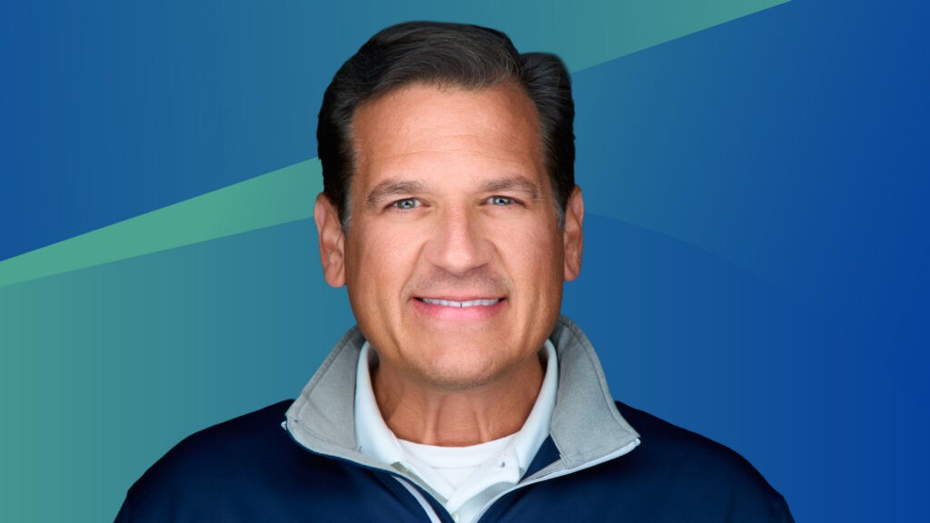 Paul Yater is pictured smiling in front of a green and blue abstract background. He has short cropped grown hair, white skin, light eyes, and wears a collared zip up over a collared white shirt.