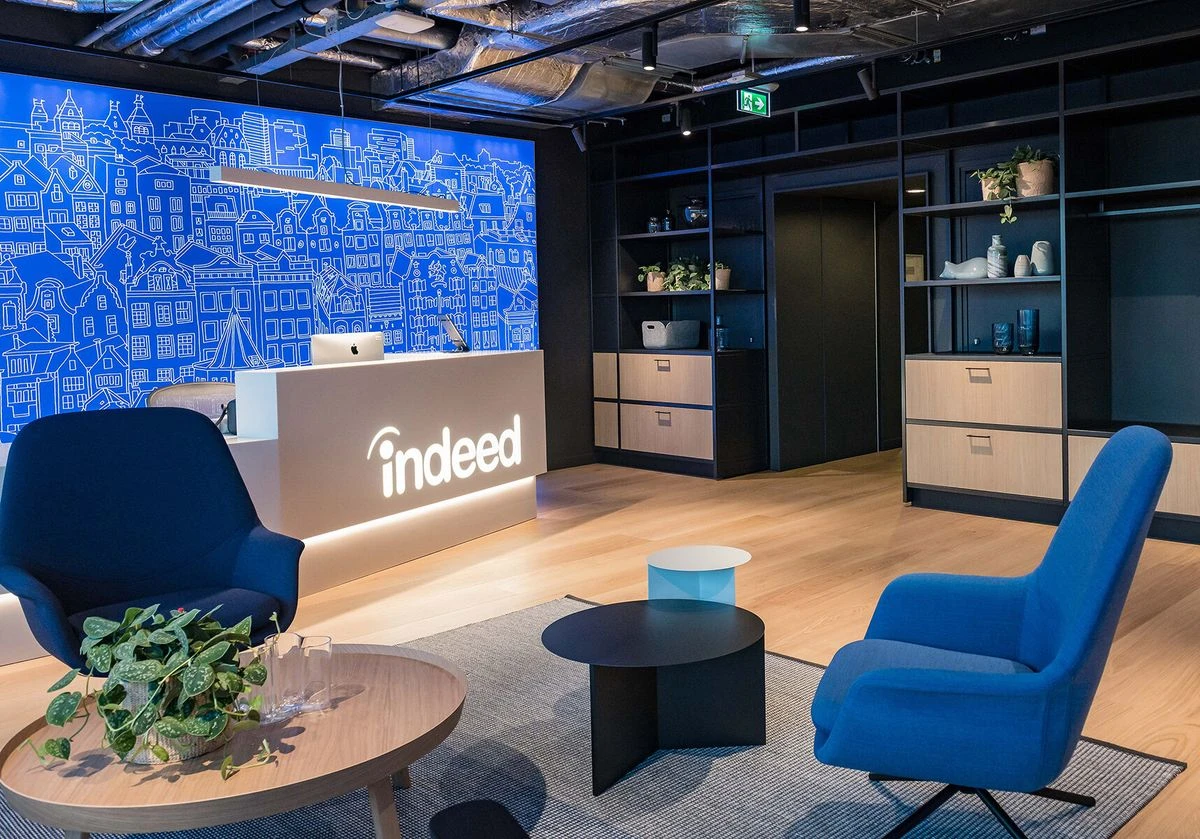A modern office reception and lounge area. A prominent white reception desk on the left features a large, illuminated Indeed logo and a glowing blue city outline mural behind it.
