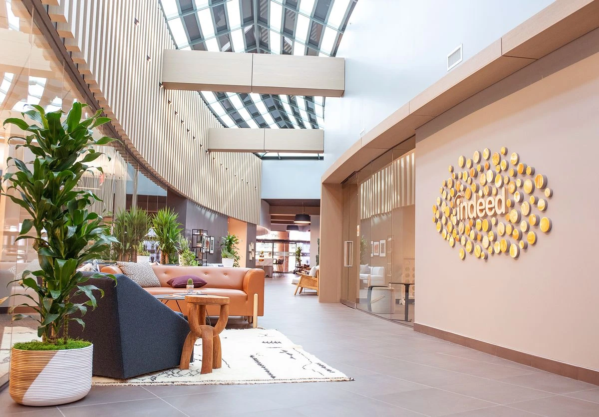A bright, modern office atrium and lobby for Indeed. The space features a large feature wall on the right with a golden oval-shaped wall art installation and the Indeed logo.