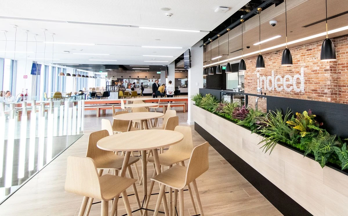 A spacious, modern office cafeteria. On the far-right, an exposed red brick wall features the Indeed logo painted in white.