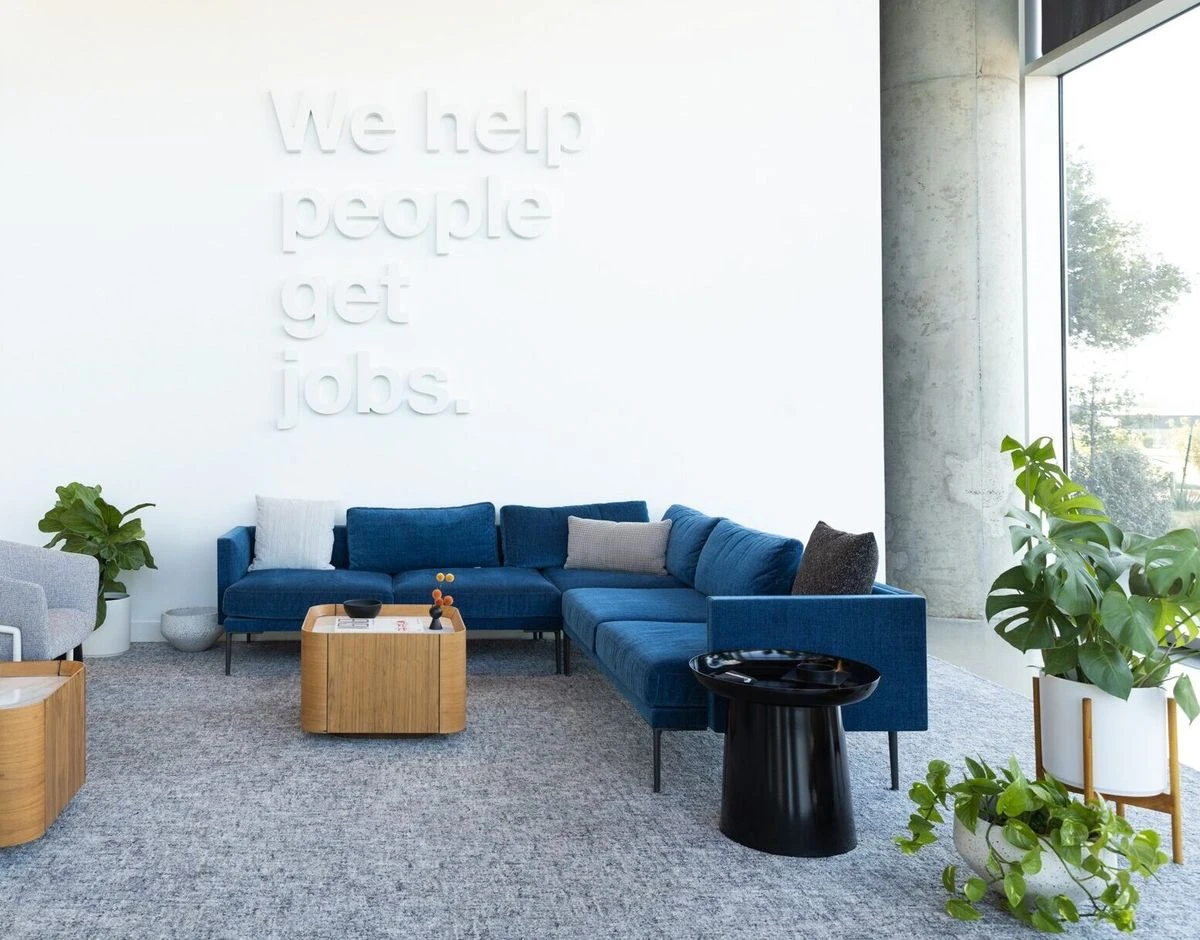 A modern office lounge area, centered on a large white wall are large, dimensional white letters that read, We help people get jobs.