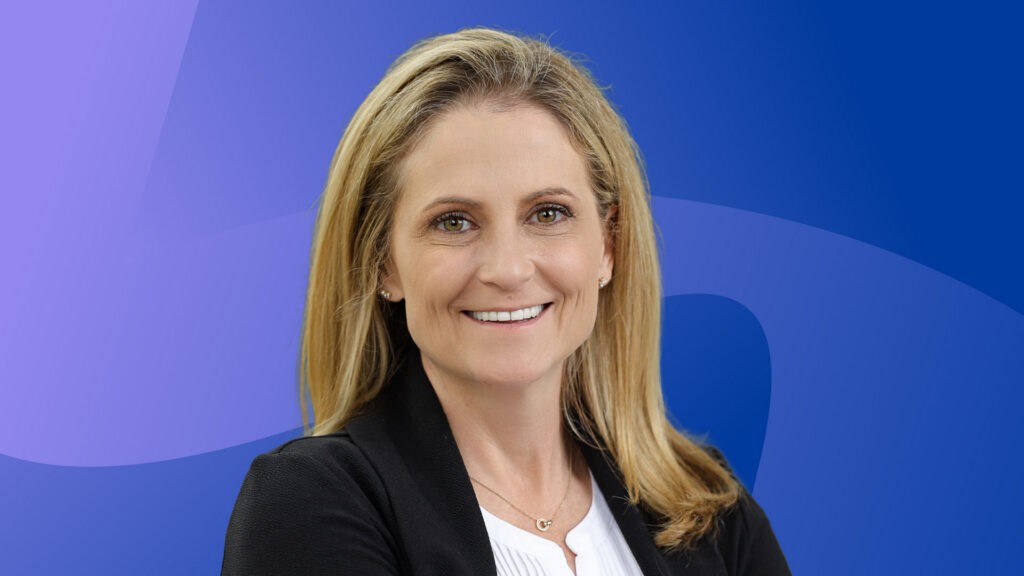 Laura Ullrich is a blonde woman with white skin wearing a white blouse and black blazer. Her headshot is in front of an indigo background with light purple swirls on it.