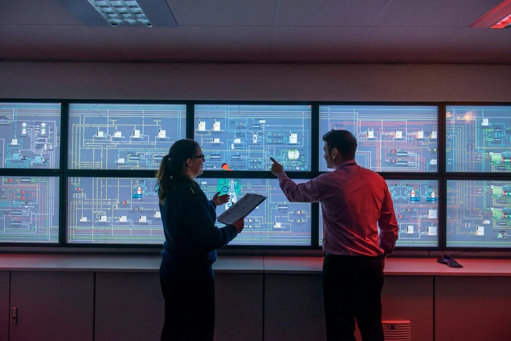 Two operators review system schematics in a control room, discussing readings across a wall of monitors displaying piping, fuel, and cooling diagrams.