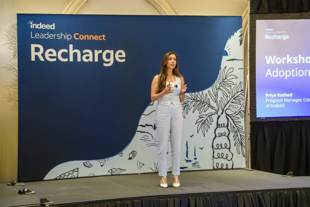 Priya Rathod on stage at the Indeed Leadership Connect Recharge stage with two large projector screens behind her introducing the agenda for the day. She is speaking and gesturing to a crowd of talent leaders gathered at round tables around the room.
