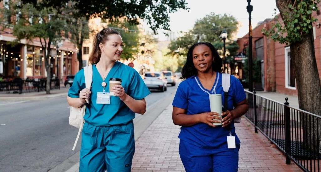 two nurses in blue scrubs walk down a street talking while carrying coffee and backpacks