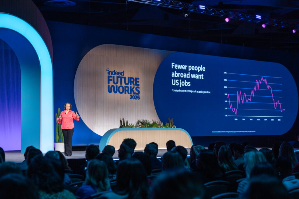 Indeed Chief Economist Svenja Gudell stands on the FutureWorks stage. She is wearing a bright pink collared shirt and black dress pants, and has short brown hair and glasses. She is speaking in front of a screen featuring a graph from Indeed Hiring Lab that reads “Fewer people abroad want U.S. jobs.”
