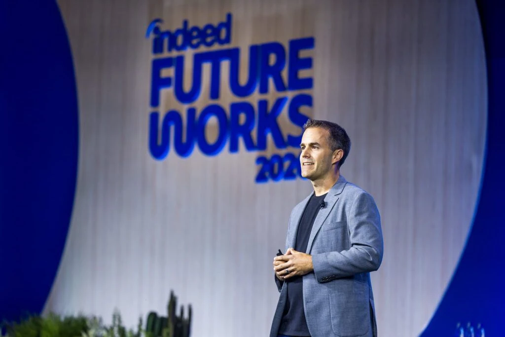 Indeed VP of Product John Fox speaks on stage in front of a backdrop reading Indeed FutureWorks 2026 in blue rounded font. He is a white man with short silver hair wearing a blue-grey suit.
