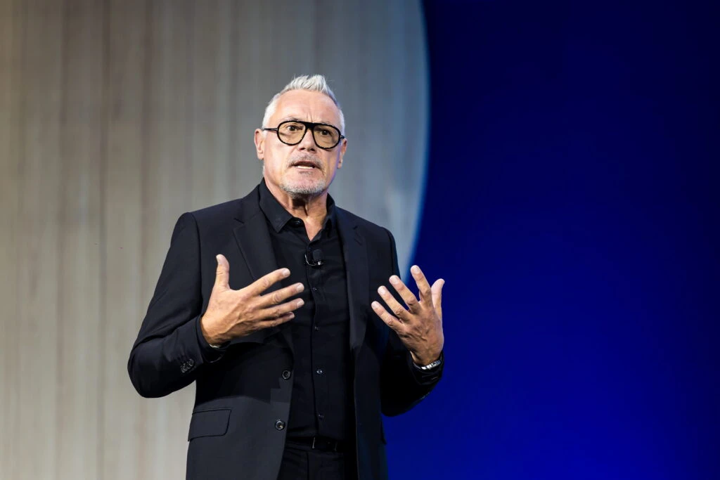 Indeed CMO James Whitemore gestures with his hands while speaking on stage in front of a bright blue backdrop. He has silver cropped hair, a goatee, dark-rimmed Aviator shaped glasses and is wearing a dark suit and button-up.
