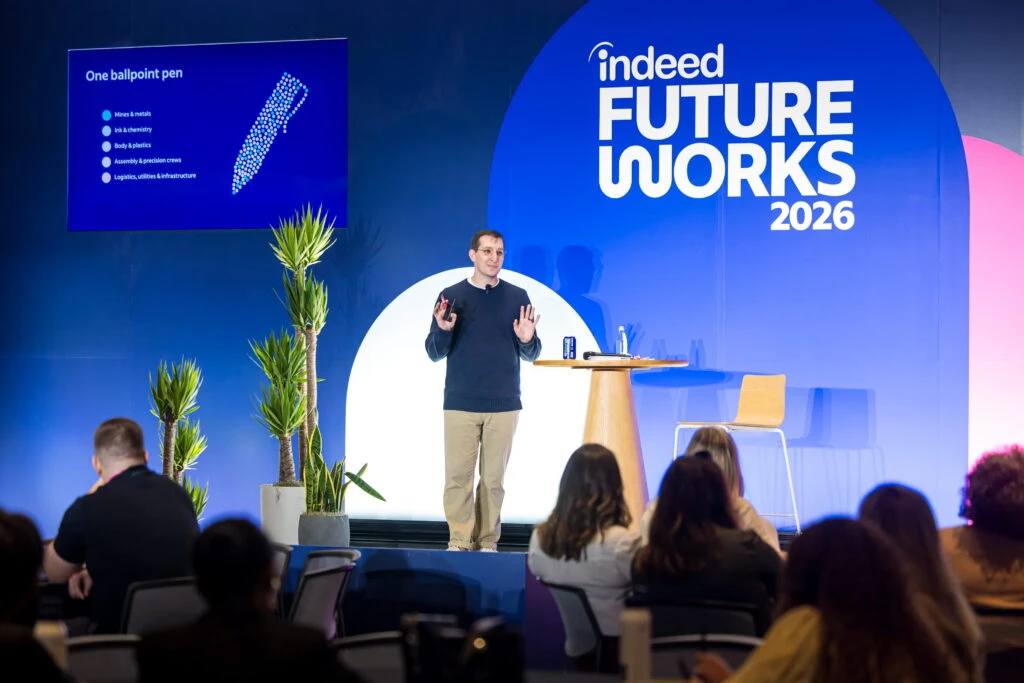 Indeed Senior Talent Strategy Advisor Kyle M.K. stands on a smaller stage decorated with desert plants and a small wooden table. He is a white man with short brown hair wearing a blue long-sleeved shirt and khakis. He is speaking to the crowd and gesturing with his hands
