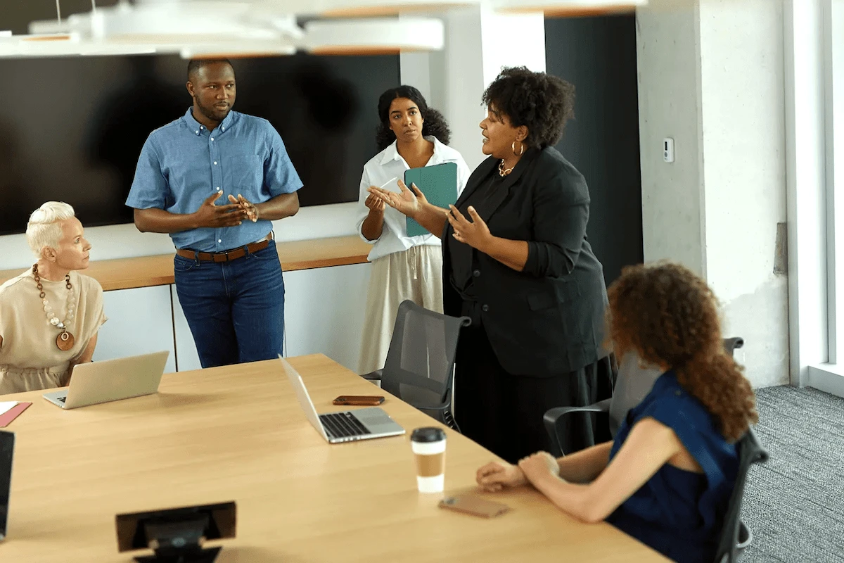 A group of five people in a modern office setting, two of them appear to be giving a presentation while the other two are seated at a wooden conference table with laptops and a coffee cup in front of them. They all seem engaged in a discussion. The room has a bright atmosphere with natural light streaming in from the side window.
