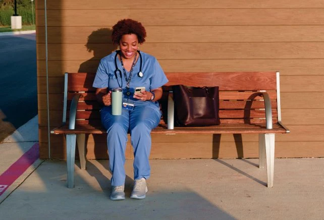 Woman in blue scrubs sitting on bench outside and looking at her phone