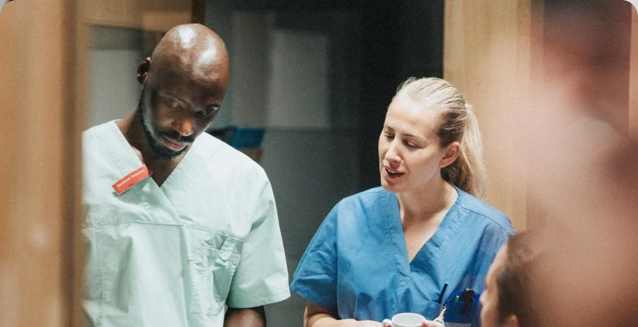 Female nurse in blue scrubs and male nurse in greens scrubs speaking to one another.