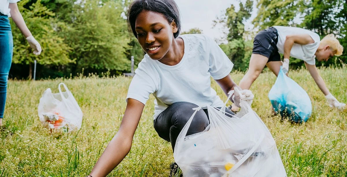 Plusieurs personnes, dont une femme de couleur souriante au centre, réduisent leur empreinte environnementale en ramassant les déchets dans un espace naturel