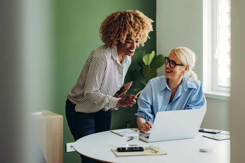 Zwei Frauen sprechen, über einen Laptop gebeugt, über Business-Themen. Sie sind beide positiv gestimmt und tauschen sich lebendig aus.