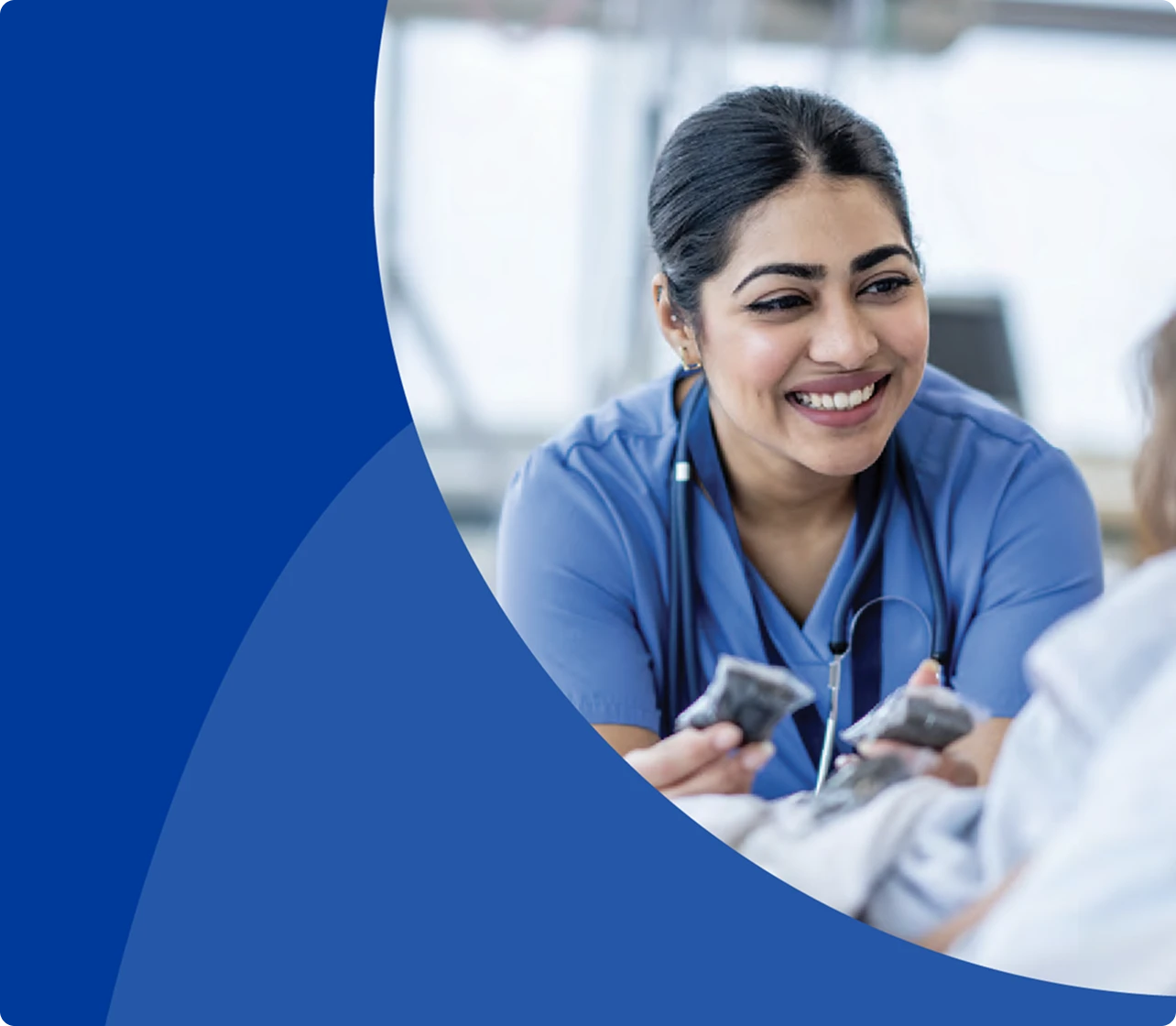 A smiling female healthcare worker in blue scrubs leans towards a patient, with a large blue graphic element on the left side of the image.