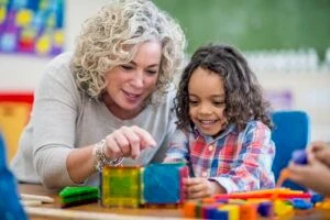 Ein Junge sitzt an seinem Schreibtisch und lächelt, während seine Vorschullehrerin auf die bunten Bauklötze vor ihm zeigt.