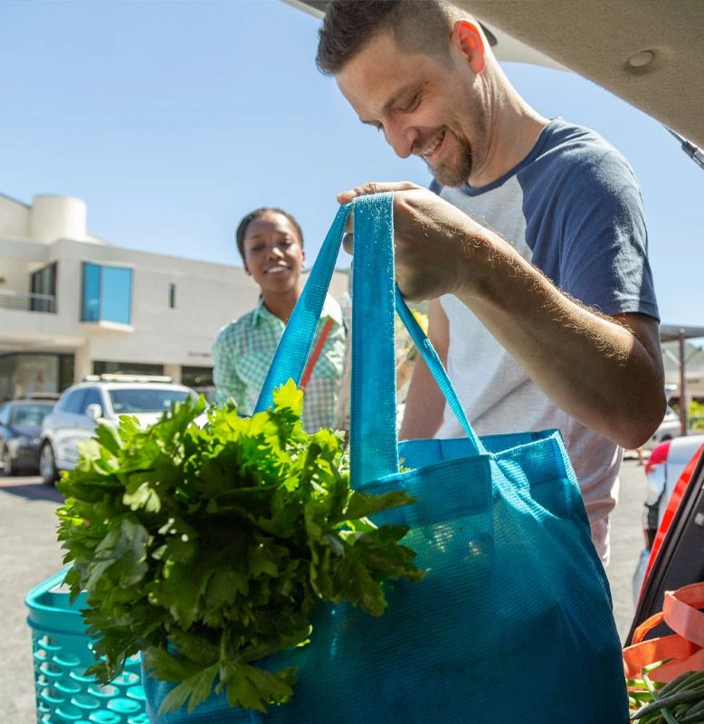 Un homme blanc range un sac réutilisable rempli de légumes verts dans sa voiture et une femme de couleur sourit en le regardant faire