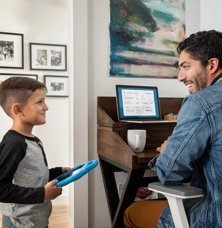 A smiling father working in his home office is approached by his child who is holding their tablet