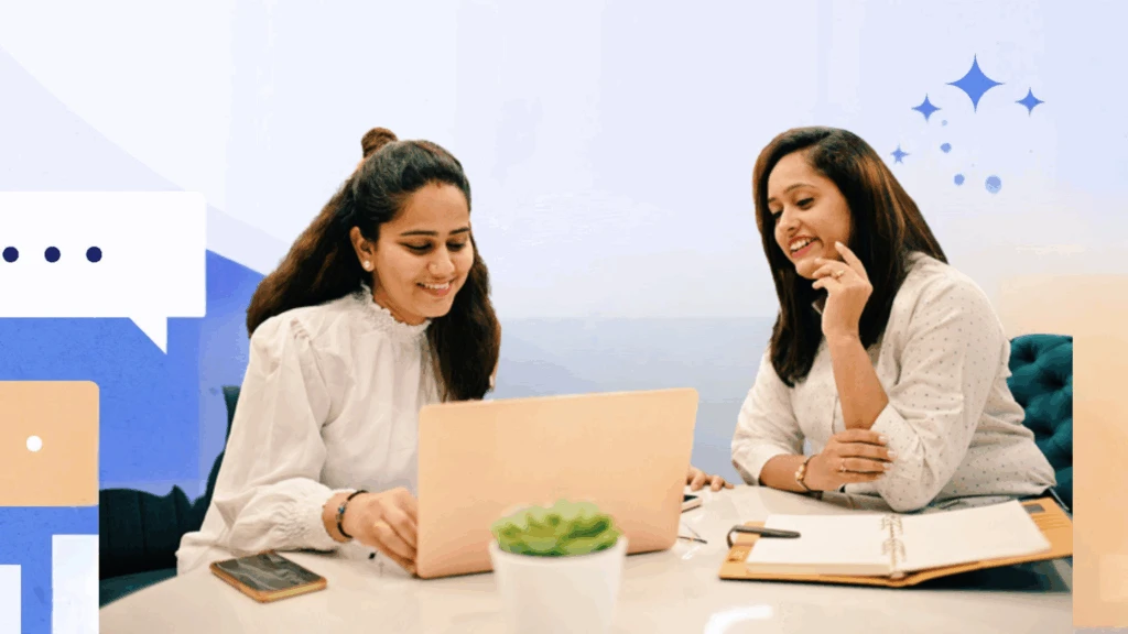Two women have an informal meeting at a table. They are looking at a laptop screen and smiling.