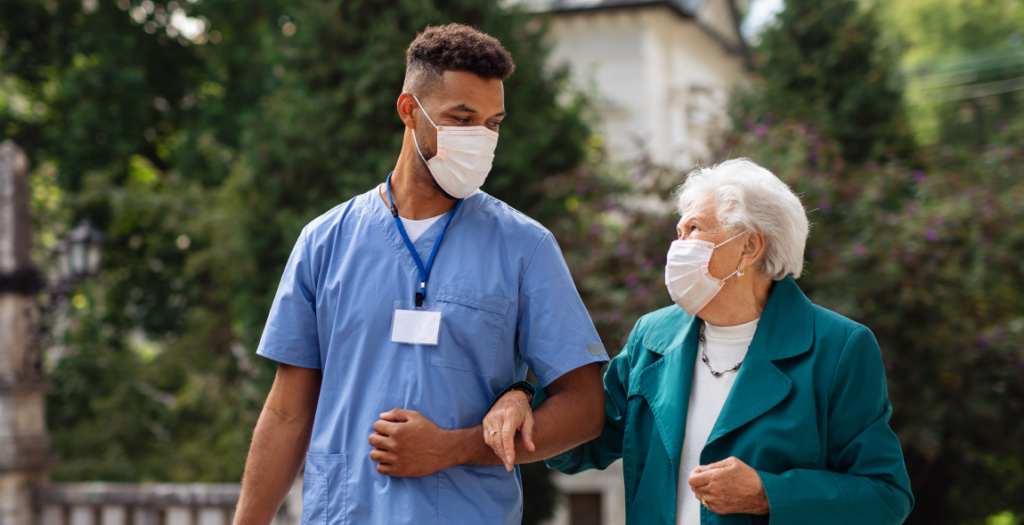 Senior woman and caregiver with face masks outdoors on a walk in park, talking, coronavirus concept.