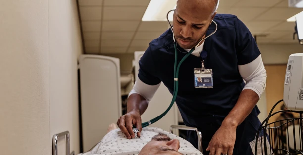 Male nurse in dark blue scrubs using a stethoscope on male patient