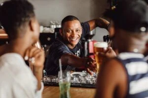 Smiling bartender reaching to give or take beer from customers out of focus in foreground
