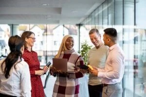 Photo of a group of business professionals in a meeting room. They are standing holding laptops/digital tablets and smiling while discussing business together.