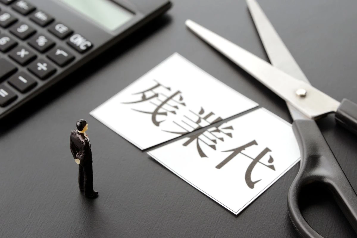 机の上に置かれた電卓、ハサミ、人形、破かれた状態の「残業代」と書かれた紙