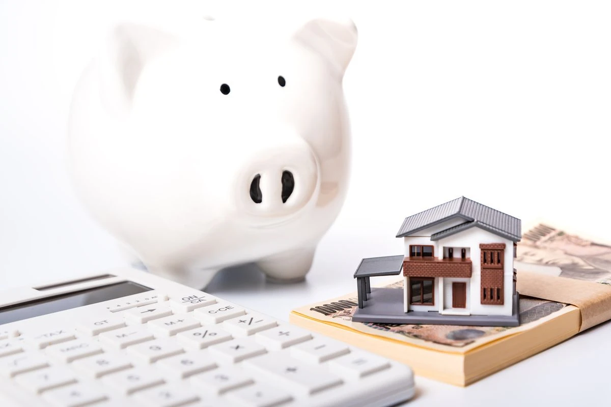 机の上に乗った電卓、お金、家の模型、豚の貯金箱のイメージ