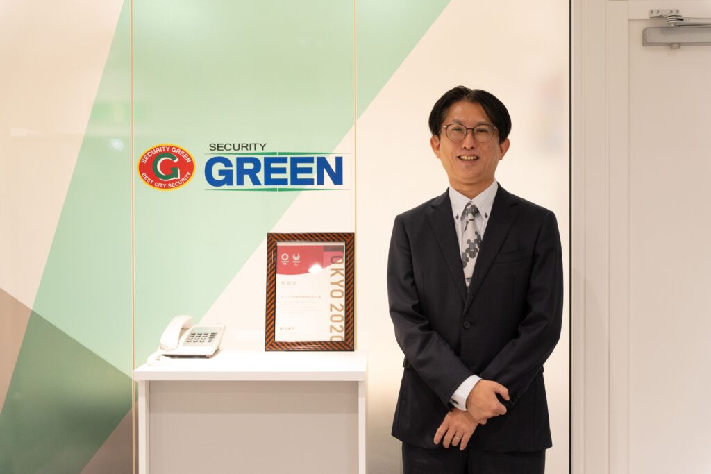 グリーン警備保障株式会社のロゴ前で微笑む山崎章司氏