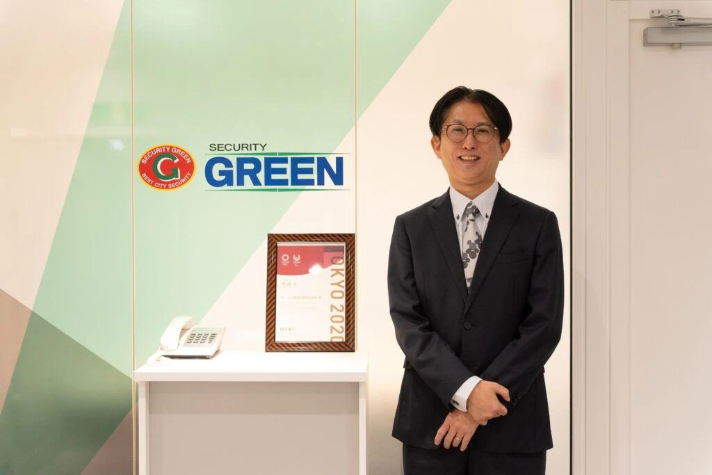 グリーン警備保障株式会社のロゴ前で微笑む山崎章司氏