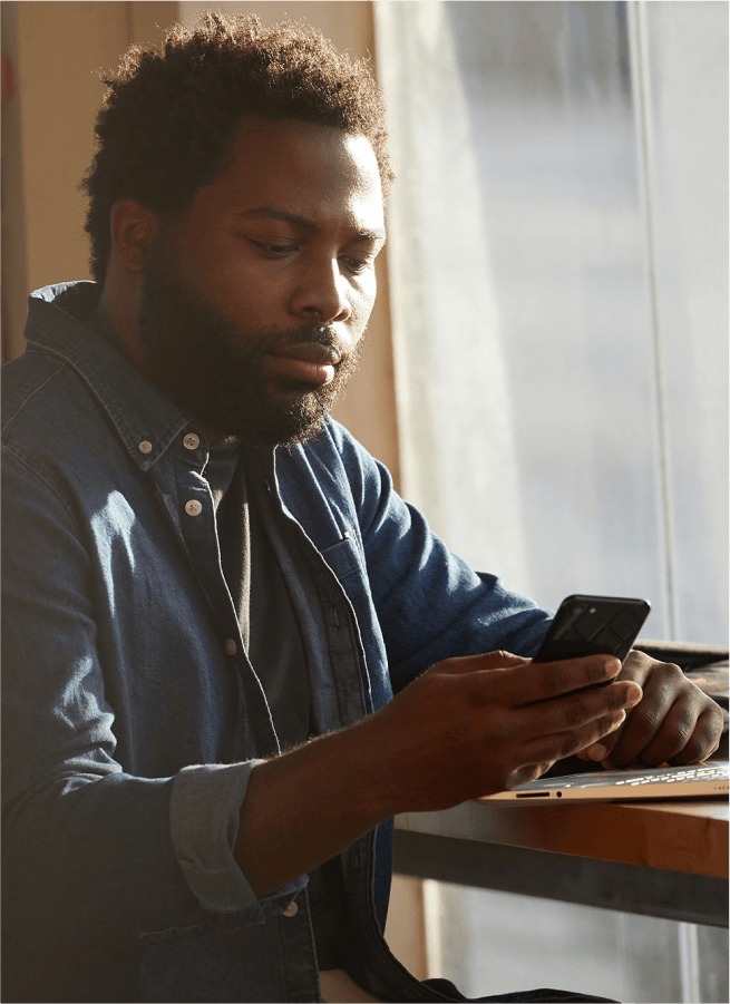 A man sitting in front of a laptop at a coffeeshop checking his phone.
