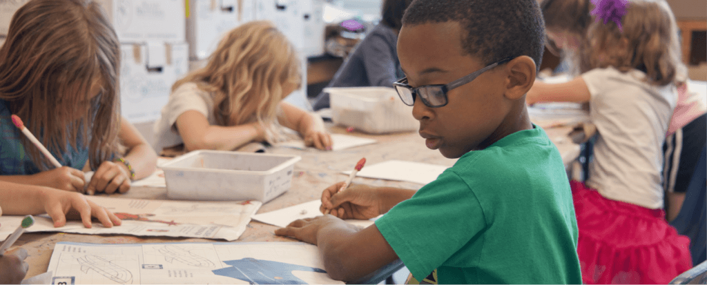 Little boy writing at a desk with classmates