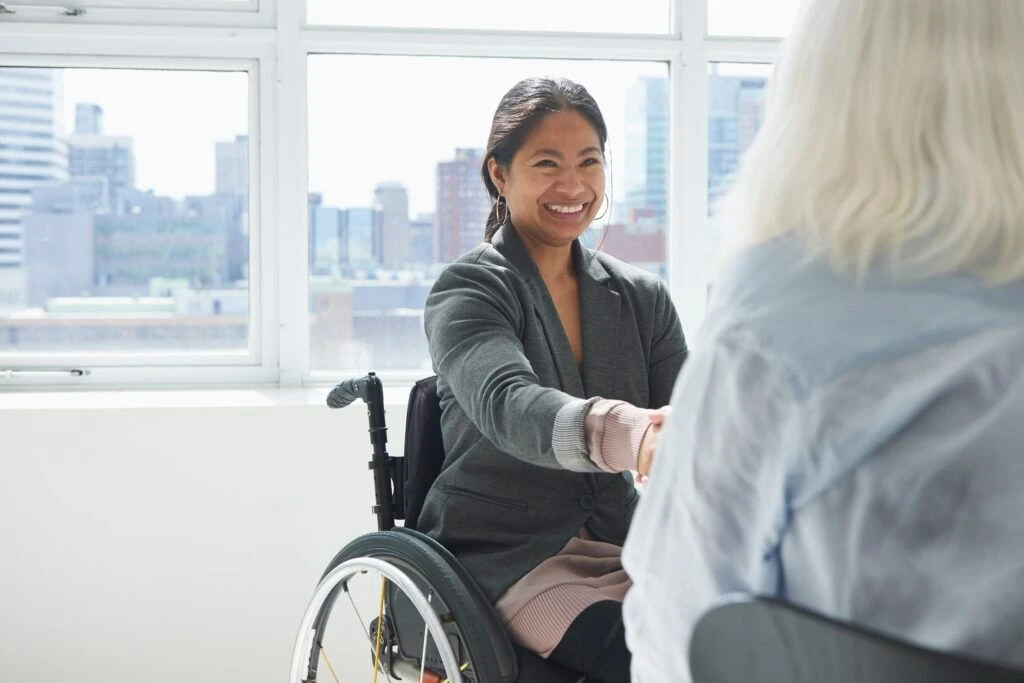 A businesswoman sitting on a wheelchair gleefully shakes another womans hand in an office setting.