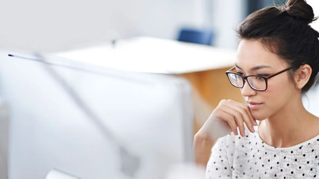 A woman with glasses pensively stares at her computer moniter.