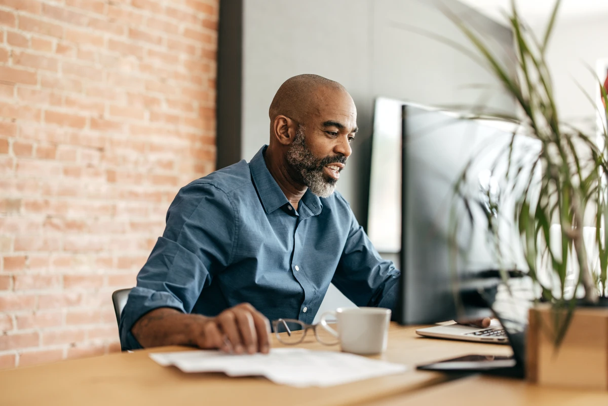 A man sitting at a desk and working on a computer while smiling. He’s in a modern office with a brick wall background and wearing a blue button-down shirt.