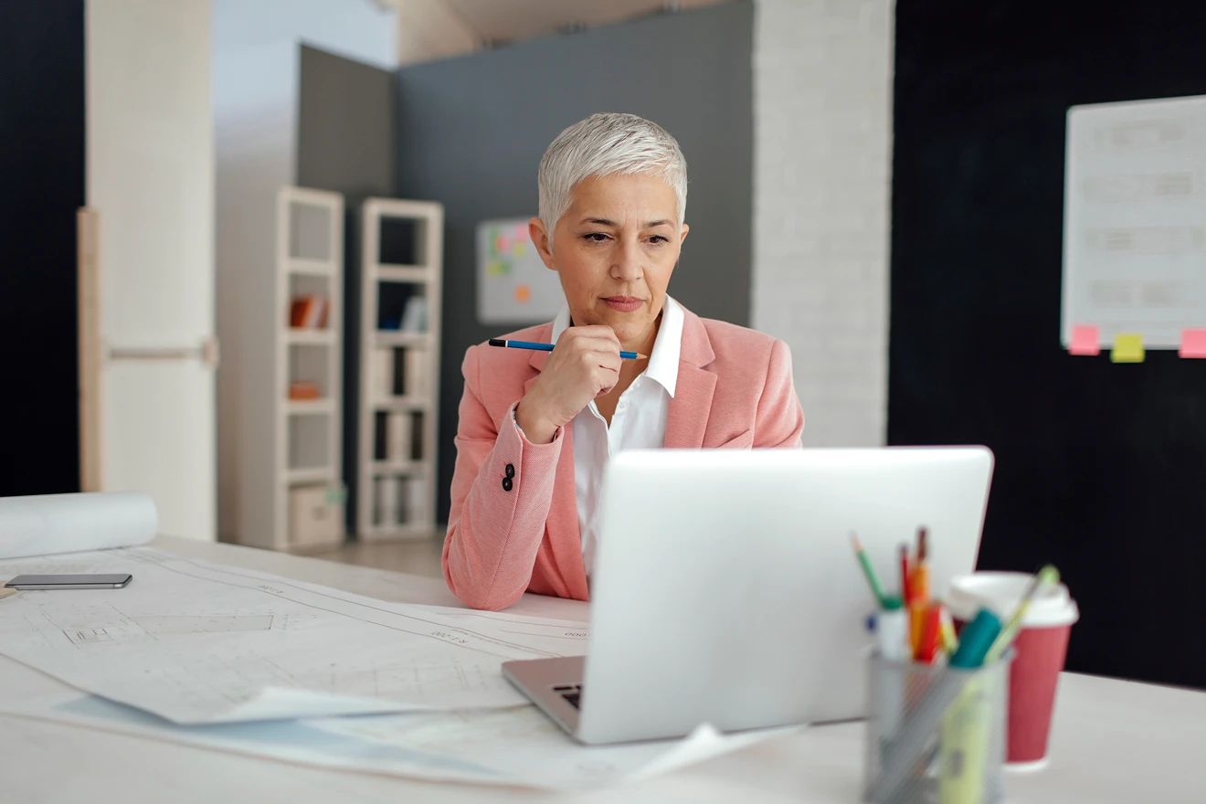 Woman holding pen, looking at laptop thoughtfully