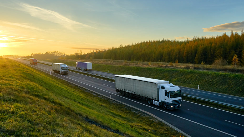 Trucks driving on the asphalt expressway between green meadows and larch forest in autumn landscape at sunset.