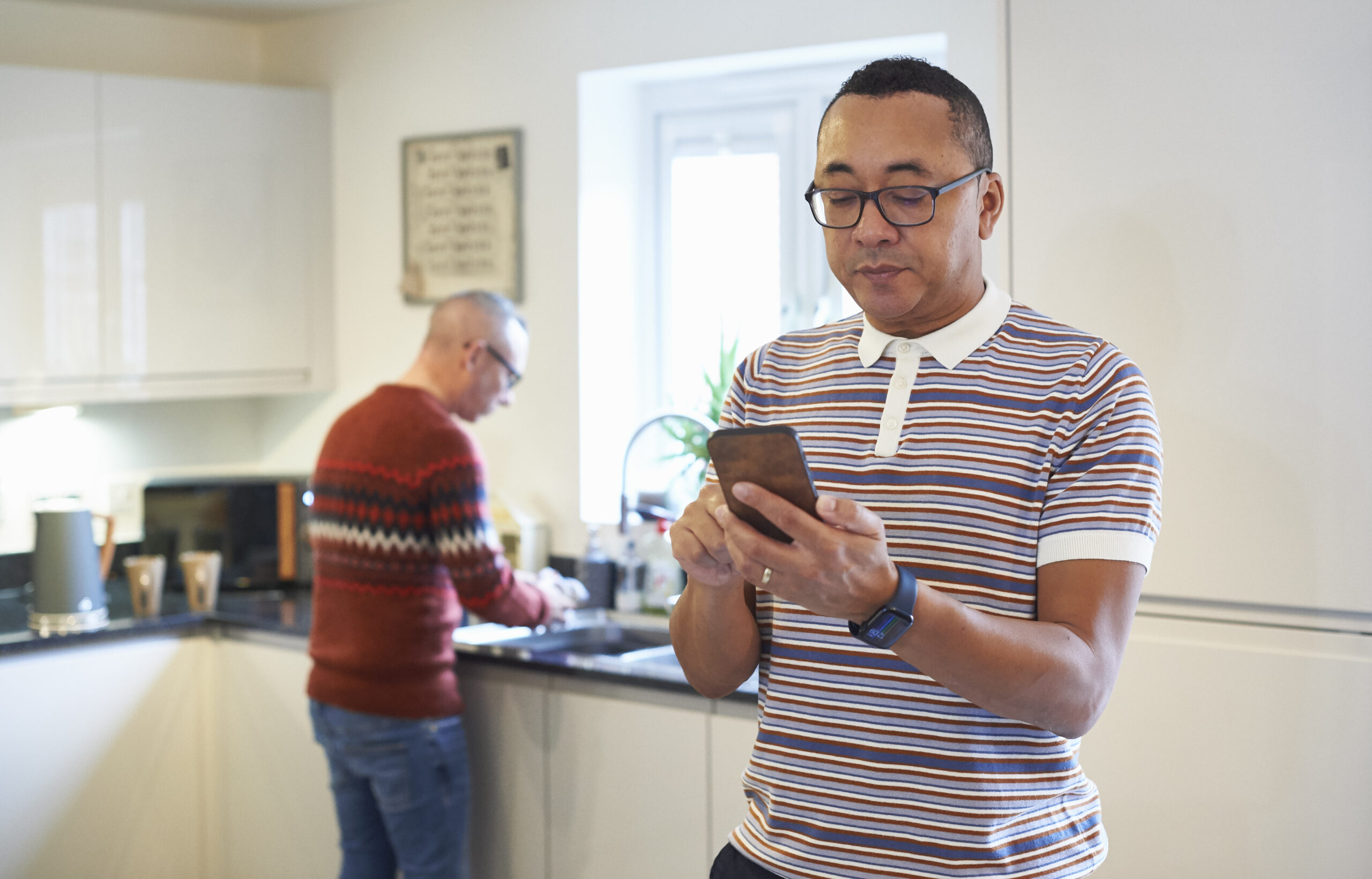 Person using smartphone in the kitchen and looking pensively at it while partner washes dishes in the background