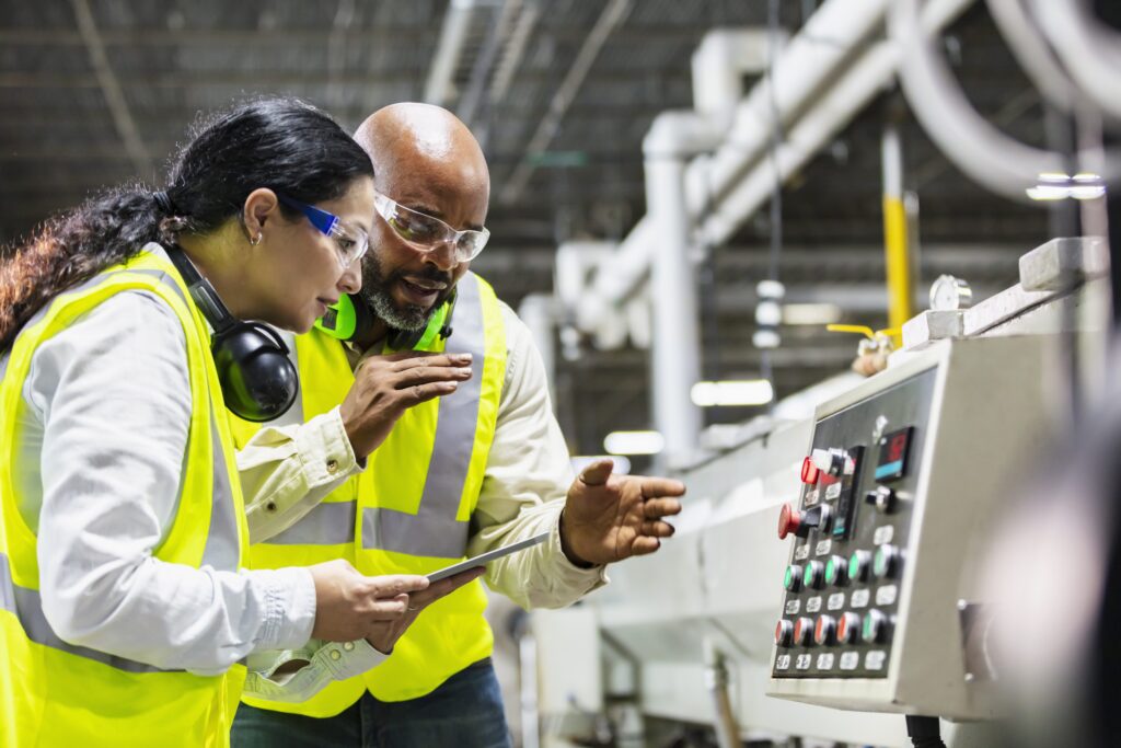 Two workers working in a plastics factory, standing on the factory floor, looking at the control panel of one of the machines.