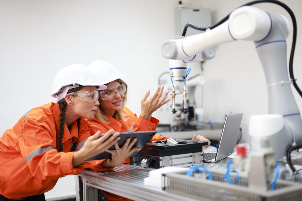 Two mechatronics engineers wearing hard hats and orange jumpsuits are working on a robotic arm in a workshop