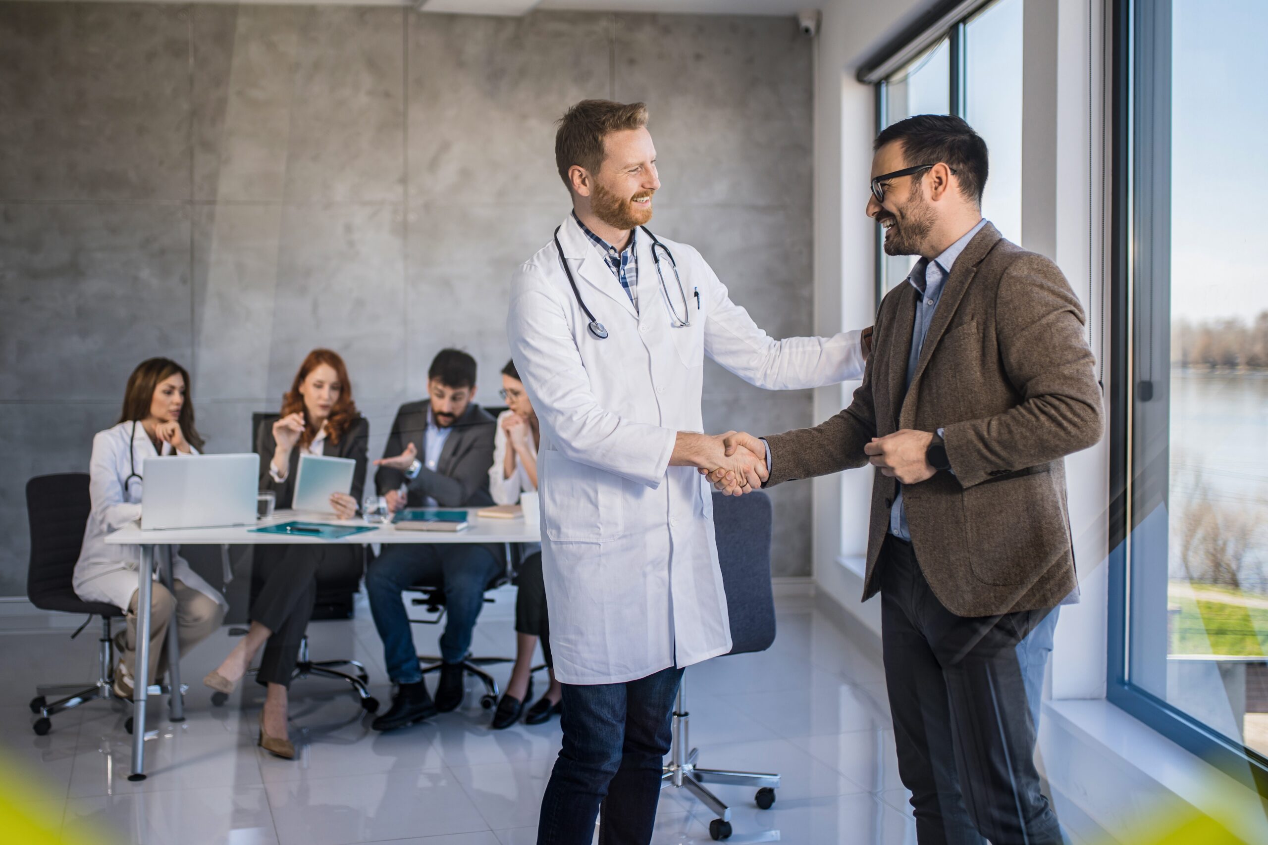 Young doctor and businessman making an agreement and shaking hands in the office. Other people are working in the background.