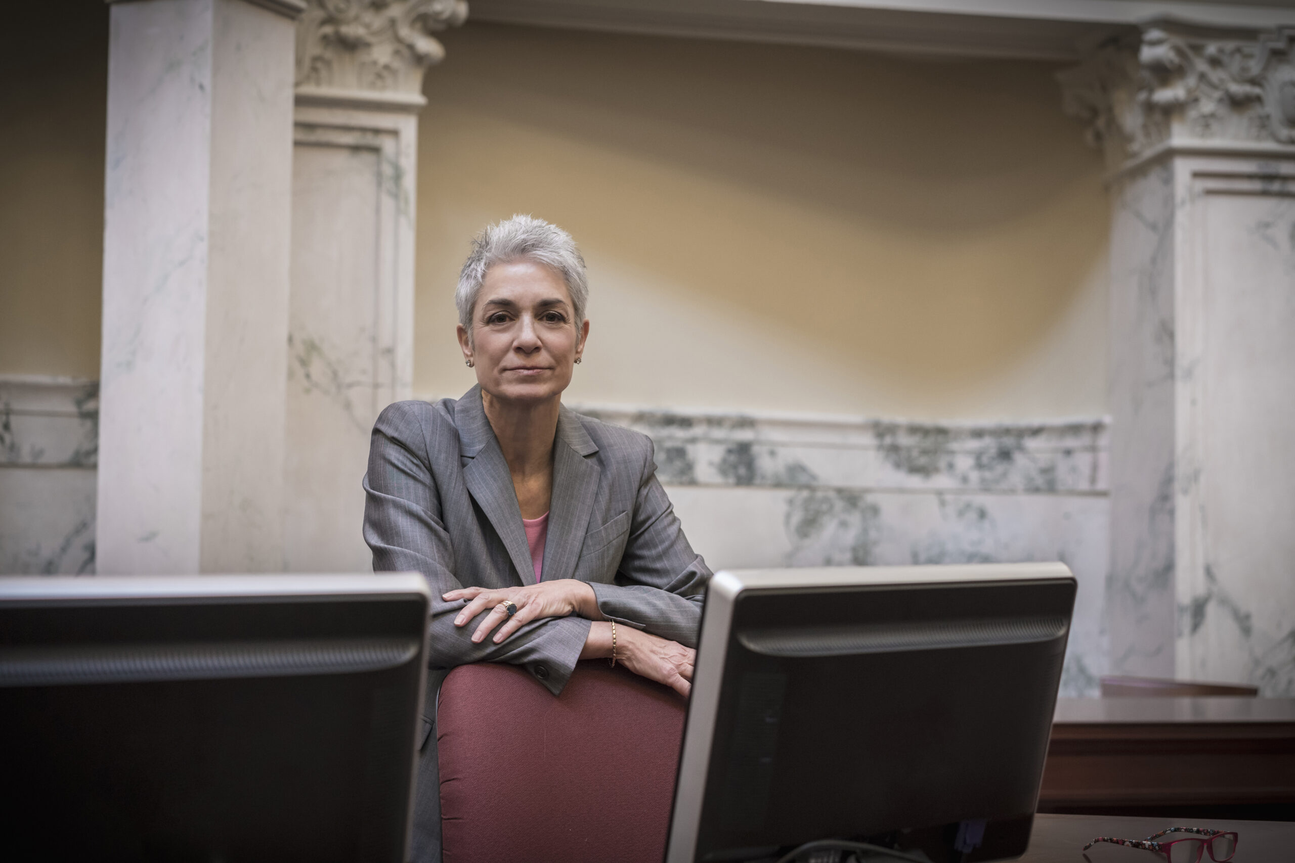 Person leaning against chair inside government building, they are wearing a suit leaning against a chair, with marble columns in the background