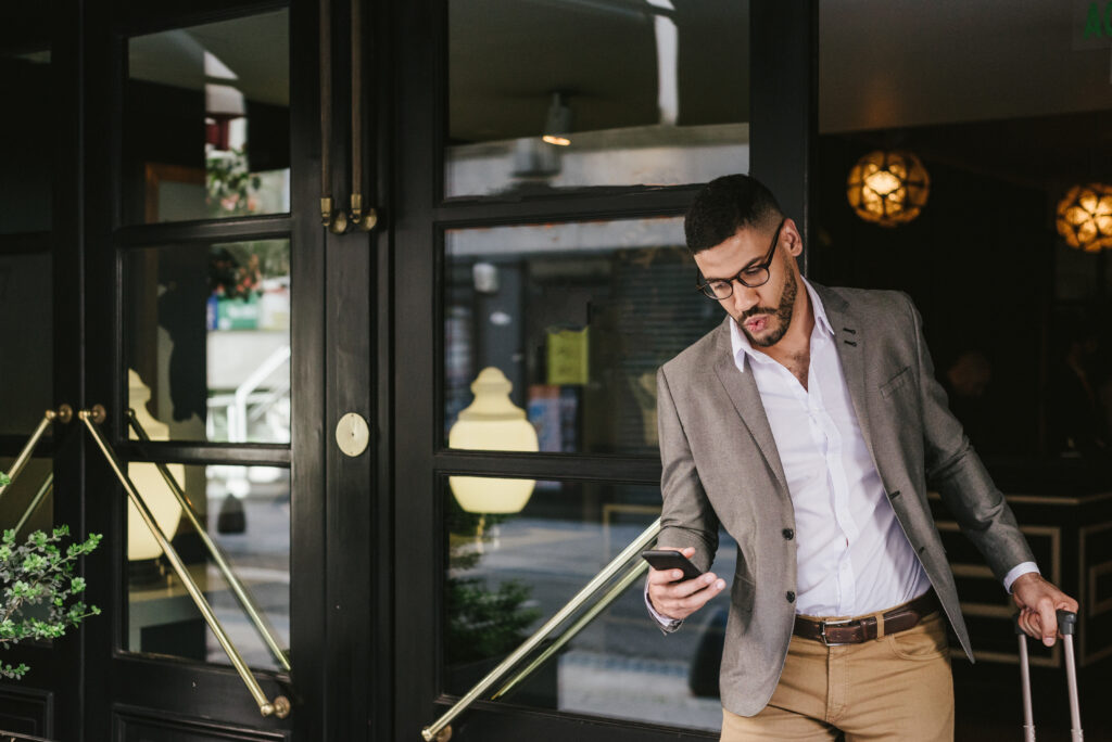 Business person leaving hotel looking at his phone and holding his suitcase