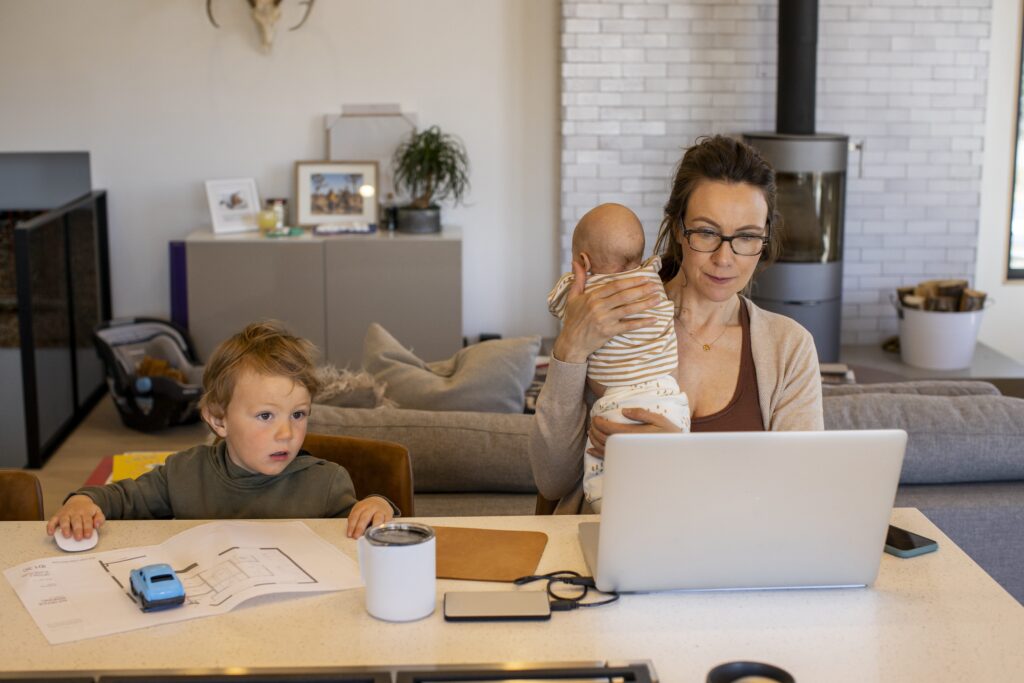 Parent carrying a baby and babysitting a toddler while using a computer at home office