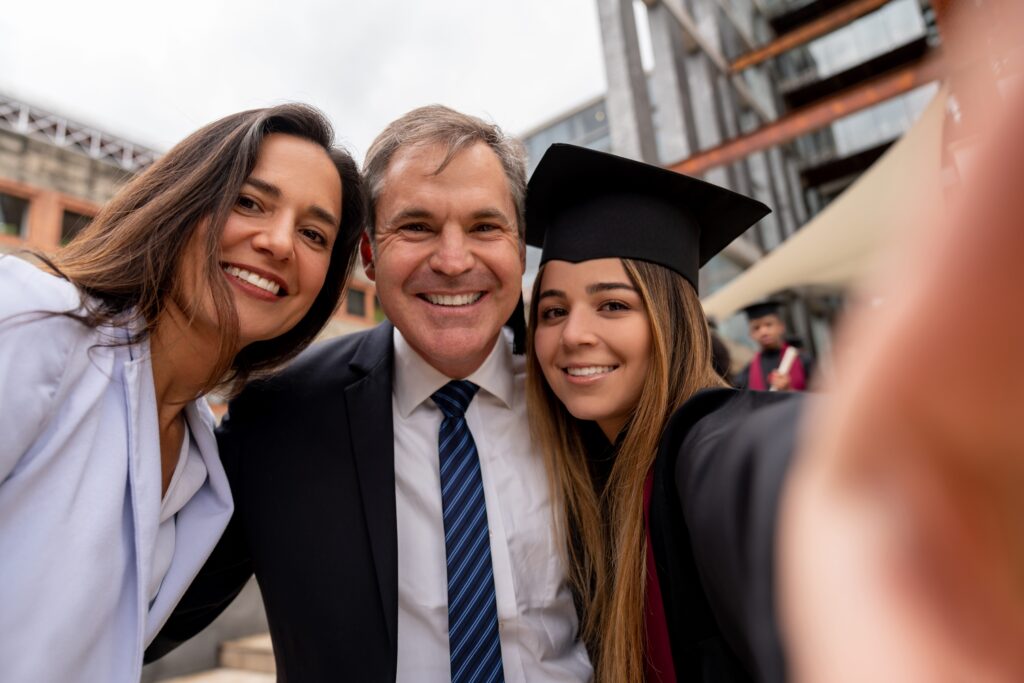 A smiling college graduate wearing a black cap and gown with a burgundy stole takes a selfie with two proud family members outdoors on a university campus. Another graduate is visible in the background.
