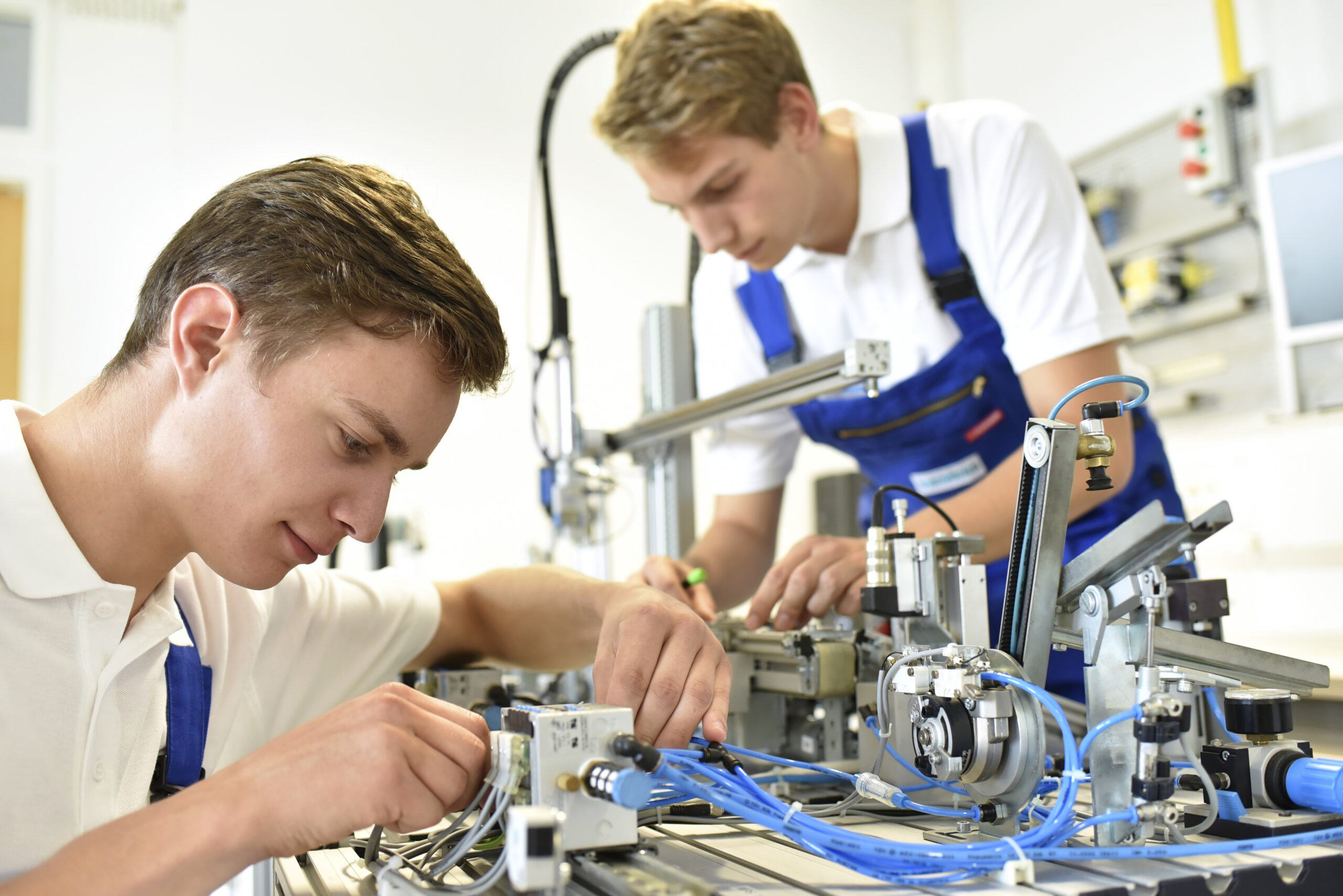 Two young technicians wearing white shirts and blue denim overalls lean over a workbench assembling a complex piece of industrial machinery. The foreground technician carefully handles wiring and blue pneumatic tubing, while his colleague in the background writes notes or adjusts a component. Metal rails, mechanical actuators, and various cables cover the work surface. A bright industrial workspace is visible in the background.