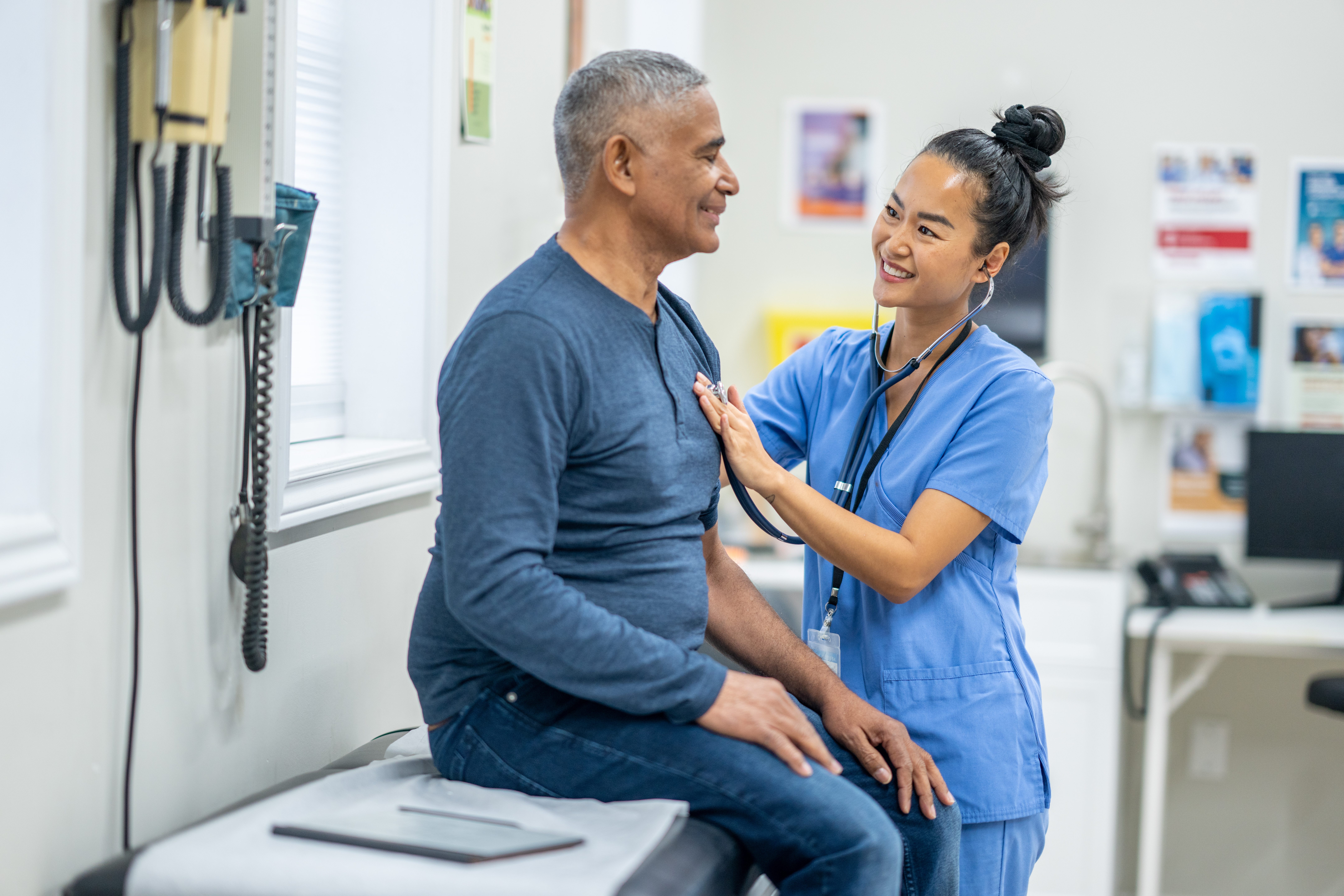 A smiling nurse in blue scrubs uses a stethoscope to listen to the chest of a middle-aged male patient seated on an exam table in a medical office. Both individuals are smiling and appear at ease. Medical equipment, including a blood pressure cuff, is mounted on the wall in the background, along with health information posters. The setting suggests a primary care or general practice clinic.