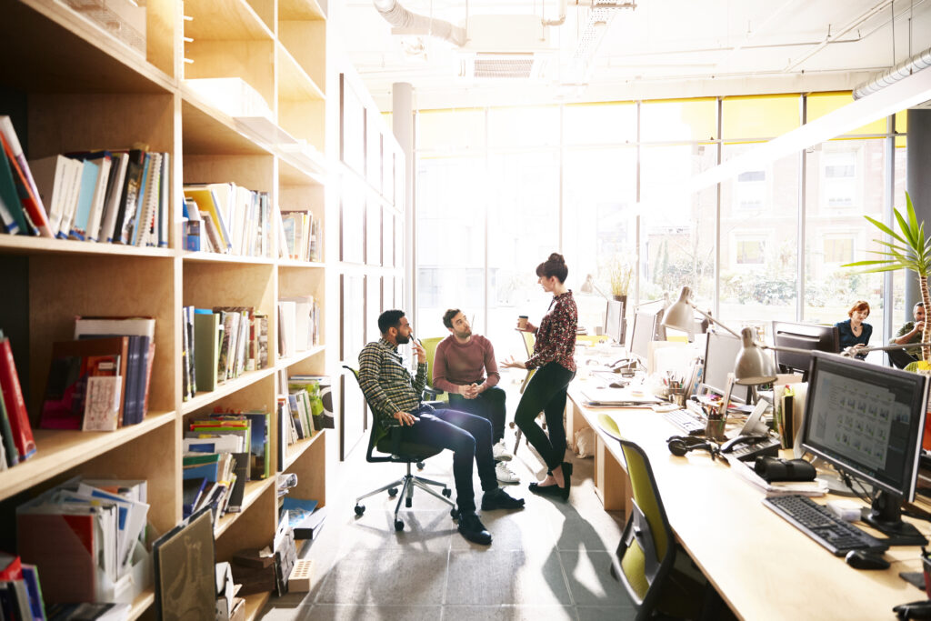 Three people having a casual conversation in a sunlit open-plan office. Two are seated while one stands holding a coffee cup. Bookshelves line the left wall and other colleagues work at desks in the background.